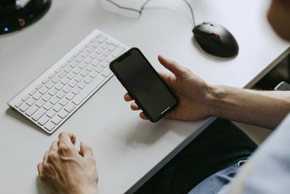 Mann sitzt am Schreibtisch und hält Smartphone in der Hand.