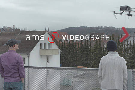 Zwei Personen sind von hinten auf einem Hausdach stehend zu sehen. Über ihnen fliegt eine Drohne und ist das ams-Videograph-Logo zu sehen